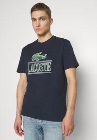 Camiseta de algodón azul marino con un gran logo de cocodrilo verde y el texto "LACOSTE" en blanco y verde, con escote redondo y mangas cortas.