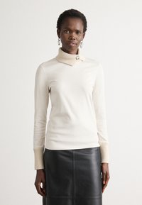 Kremasti pulover s turtleneck izrezom, s rebrastim bež rukavima i okovratnikom, ukrašen dekorativnim dugmetom. Uparen s crnom kožnom suknjom.