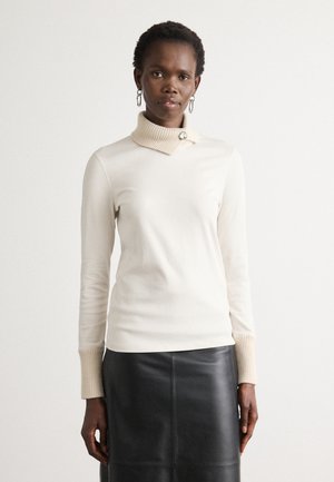 Cremfarbener Rollkragenpullover mit gerippten beigen Bündchen und Kragen, ausgestattet mit einem dekorativen Knopf. Kombiniert mit einem schwarzen Lederrock.