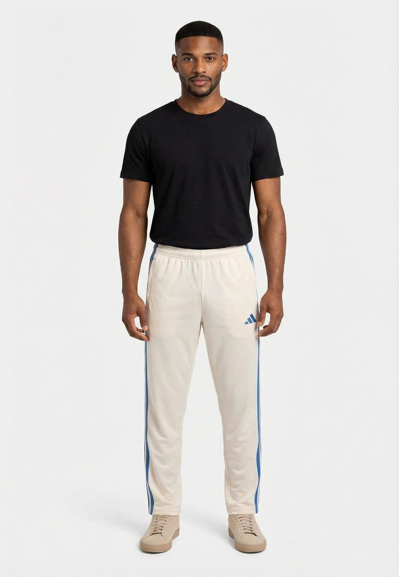 Uomo in piedi di fronte, indossa una maglietta nera, pantaloni sportivi Adidas bianchi con strisce blu e sneakers beige su sfondo bianco.