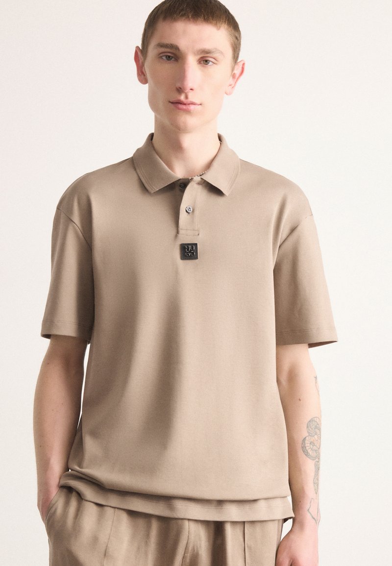 Beige polo shirt met een klassieke kraag, korte mouwen en een gestructureerd oppervlak; heeft een klein logo patch nabij de kraag.