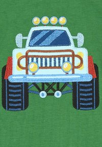 Sininen ja punainen monsterirekka, joka on brodeerattu, ja jossa on keltaiset valot, mustat renkaat ja vihreä tausta. Yksityiskohtainen etusäleikkö ja muotoilulinjat.