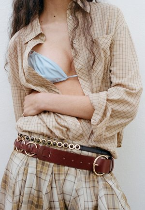 Mujer que lleva camisa de cuadros beige abierta para revelar un sujetador azul claro, combinada con falda de cuadros a juego y cinturón marrón en capas con anillas metálicas.