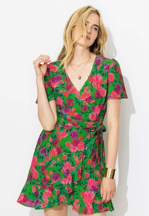 Femme portant une robe portefeuille verte à manches courtes avec un imprimé floral rose et violet, un collier en or et un bracelet manchette en or, posant sur un fond blanc.