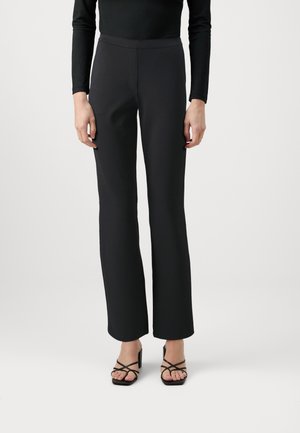 Personne portant un pantalon noir taille haute à jambes droites, un haut noir à manches longues et des sandales à talons noires à brides et bouts ouverts sur fond blanc.