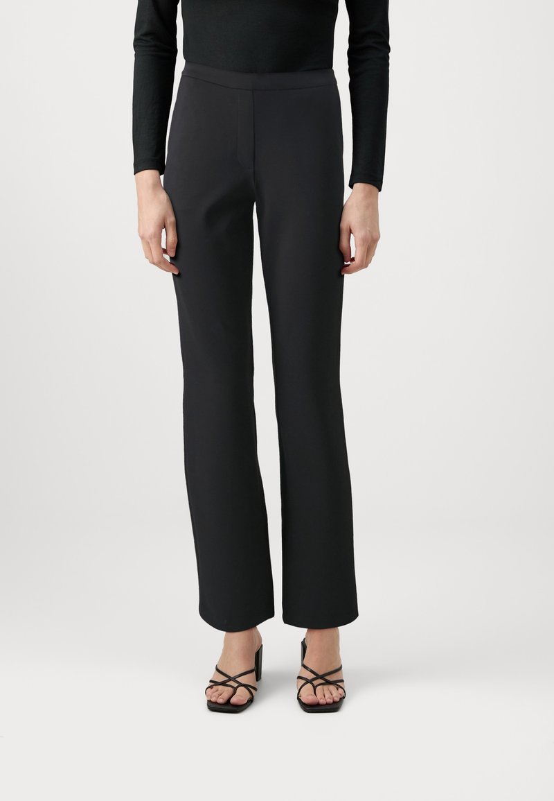 Personne portant un pantalon noir taille haute à jambes droites, un haut noir à manches longues et des sandales à talons noires à brides et bouts ouverts sur fond blanc.
