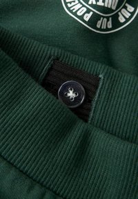 Zaļa džemperis ar ribotām aprocēm. Iekļauts skaidrs pogas un tekstūrēts melns jostasvīrs. Cirkulārs balts logotips uz auduma.