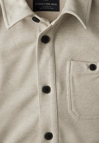 Camisa beige de botones con una tela texturizada, que presenta un bolsillo en el pecho y botones negros. La etiqueta dice "STREET ONE MEN."