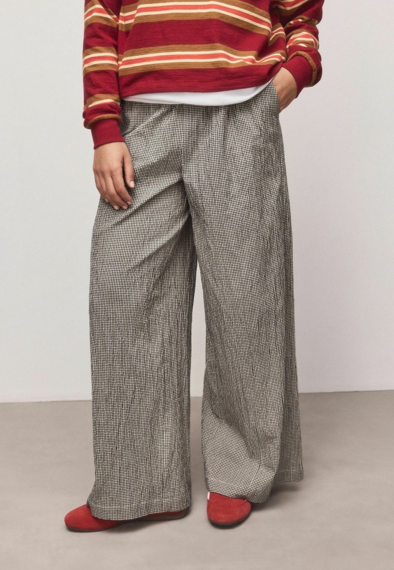Plaša stila bikses ar melnbaltu ginghema rakstu, izgatavotas no faktūras auduma. Kombinētas ar sarkaniem dzīvokļiem un svītrainu bordo krāsas topu.