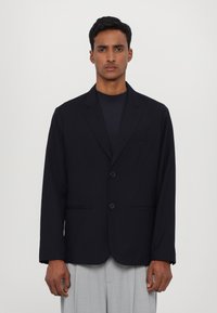 Marineblazer van gladde stof, met een notch lapel, een sluiting met twee knopen en twee zijzakken. Gecombineerd met lichtgrijze broeken.
