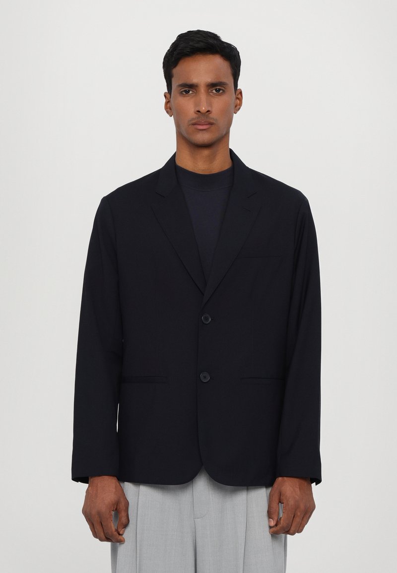 Marineblazer van gladde stof, met een notch lapel, een sluiting met twee knopen en twee zijzakken. Gecombineerd met lichtgrijze broeken.