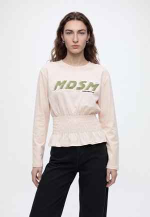 Donna con lunghi capelli ricci che indossa una maglia a maniche lunghe beige chiaro con testo verde "MDSM COLLECTIVE" e pantaloni neri su uno sfondo uniforme.
