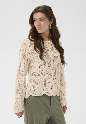 Donna con capelli lunghi castani, che indossa orecchini a cerchio dorati e un top crema all'uncinetto con bottoni abbinato a pantaloni verdi a vita alta, guarda verso sinistra.