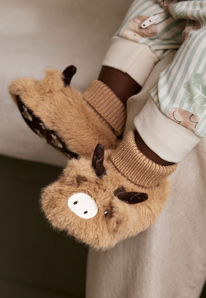 Enfant portant des chaussons bruns en fourrure, conçus comme des animaux avec de petites cornes et des museaux blancs.