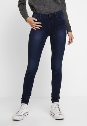 Persoon draagt donkerblauwe skinny jeans, grijze trui en witte hoge sneakers, staat met één hand op de heup.