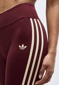 Gros plan sur une personne portant des leggings bordeaux avec le design à trois bandes Adidas beige et le logo sur la hanche, la main reposant à côté de la cuisse.