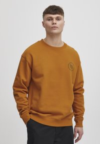 Majica mustard boje napravljena od mekog materijala. Ima okrugli izrez, rebraste manžetne i mali detalj s logotipom s lijeve strane.