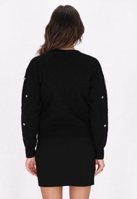 Schwarzer Pullover mit Rautenmuster, geripptem Saum und herzförmigen Nieten an den Ärmeln. Kombiniert mit einem taillierten schwarzen Rock. Strukturierte Strickmaterial.
