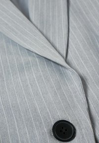 Blazer gris clair à fines rayures avec un bouton noir à quatre trous et un col à revers cranté.