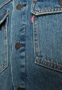 Jeansjacka med blått tyg, kopparfärgad söm, två framfickor med lock och en liten röd logotypetikett på vänster sida.