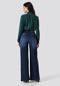 Chemise verte à manches longues associée à un jean en denim bleu taille haute et jambes larges. La chemise présente un col discret et un détail à l'arrière.