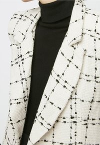 Blazer tessuto in bianco con un motivo a rete nera. Presenta spalle e rever strutturati. Indossato sopra un dolcevita nero. Tessuto testurizzato.