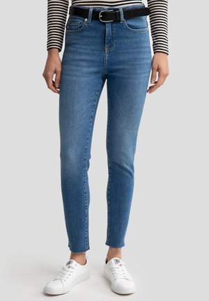 Blaue hochgeschnittene Skinny-Jeans aus Denim, mit vorderen Taschen, einem Reißverschluss und rohem Saum. Kombiniert mit weißen Sneakers.