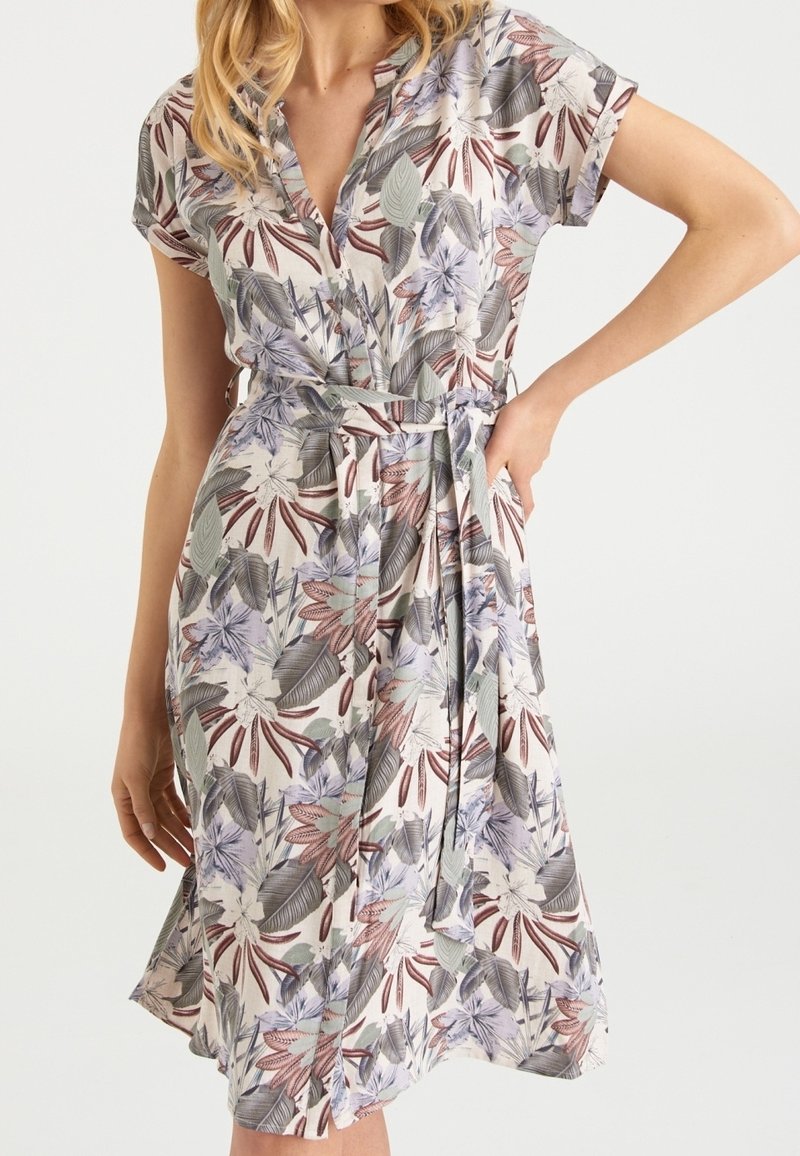 Sukienka koszulowa w kwiatowy wzór z krótkimi rękawami, paskiem w talii i guzikami z przodu, noszona przez osobę z jedną ręką na biodrze.