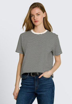 Jonge vrouw met lang haar, gekleed in een zwart-wit gestreept shirt met korte mouwen en blauwe spijkerbroek, hand in de zak, neutrale achtergrond.