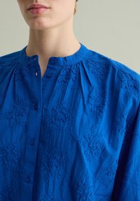 Camicia blu ricamata con scollo rotondo, chiusura con bottoni e motivi floreali. Realizzata in tessuto testurizzato con dettagli arricciati.