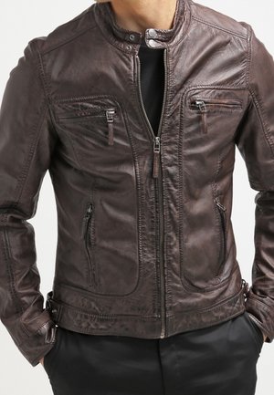 Homme portant une veste en cuir brun foncé zippée avec col montant et quatre poches zippées, les mains dans les poches du pantalon.