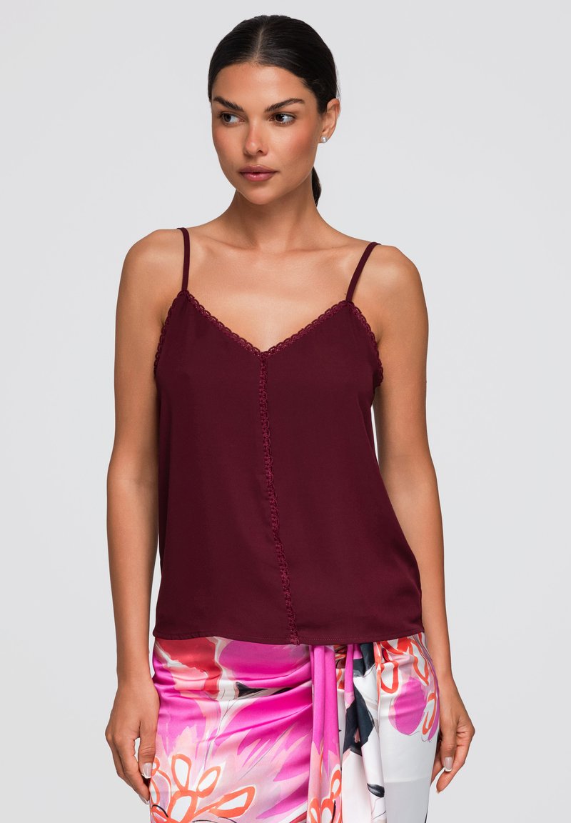 Femme aux cheveux foncés portant un haut sans manches bordeaux et une jupe portefeuille fleurie rose, blanche et orange, debout devant un fond uni.