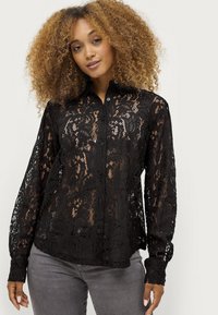 Zwarte kanten blouse met een knoopsluiting aan de voorkant, lange mouwen, bloempatronen en een doorschijnende textuur, gecombineerd met grijze jeans.