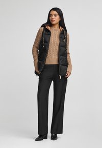 Svart pufferväst med en glänsande kopparfärgad insida över en beige kabelstickad tröja, i kombination med skräddarsydda svarta byxor och ankelboots.