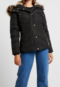 Giacca bomber nera con cappuccio foderato in pelliccia, dotata di accessori dorati, cuciture diagonali e due tasche laterali con zip.