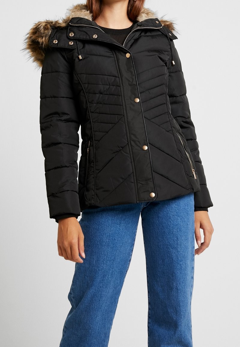 Giacca bomber nera con cappuccio foderato in pelliccia, dotata di accessori dorati, cuciture diagonali e due tasche laterali con zip.