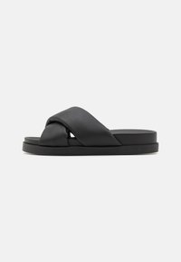 Sandales à glissière en cuir noir avec un design supérieur tordu et une semelle plate. Texture lisse avec une esthétique minimaliste et moderne.