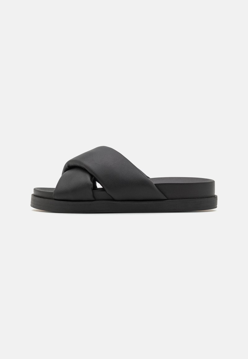 Sandales à glissière en cuir noir avec un design supérieur tordu et une semelle plate. Texture lisse avec une esthétique minimaliste et moderne.