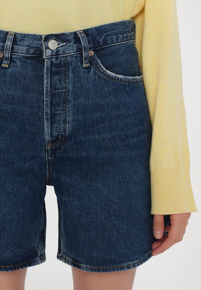 Shorts in denim a vita alta di colore blu scuro, con chiusura a bottone, due tasche frontali e orlo sfrangiato. Indossati con un maglione giallo.