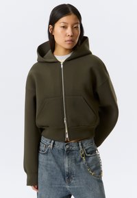 Νεαρή γυναίκα που φοράει μια σκούρα πράσινη cropped ζακέτα με φερμουάρ και μπλε τζιν με μεταλλική αλυσίδα στην δεξιά τσέπη, στέκεται μπροστά σε ένα απλό φόντο.