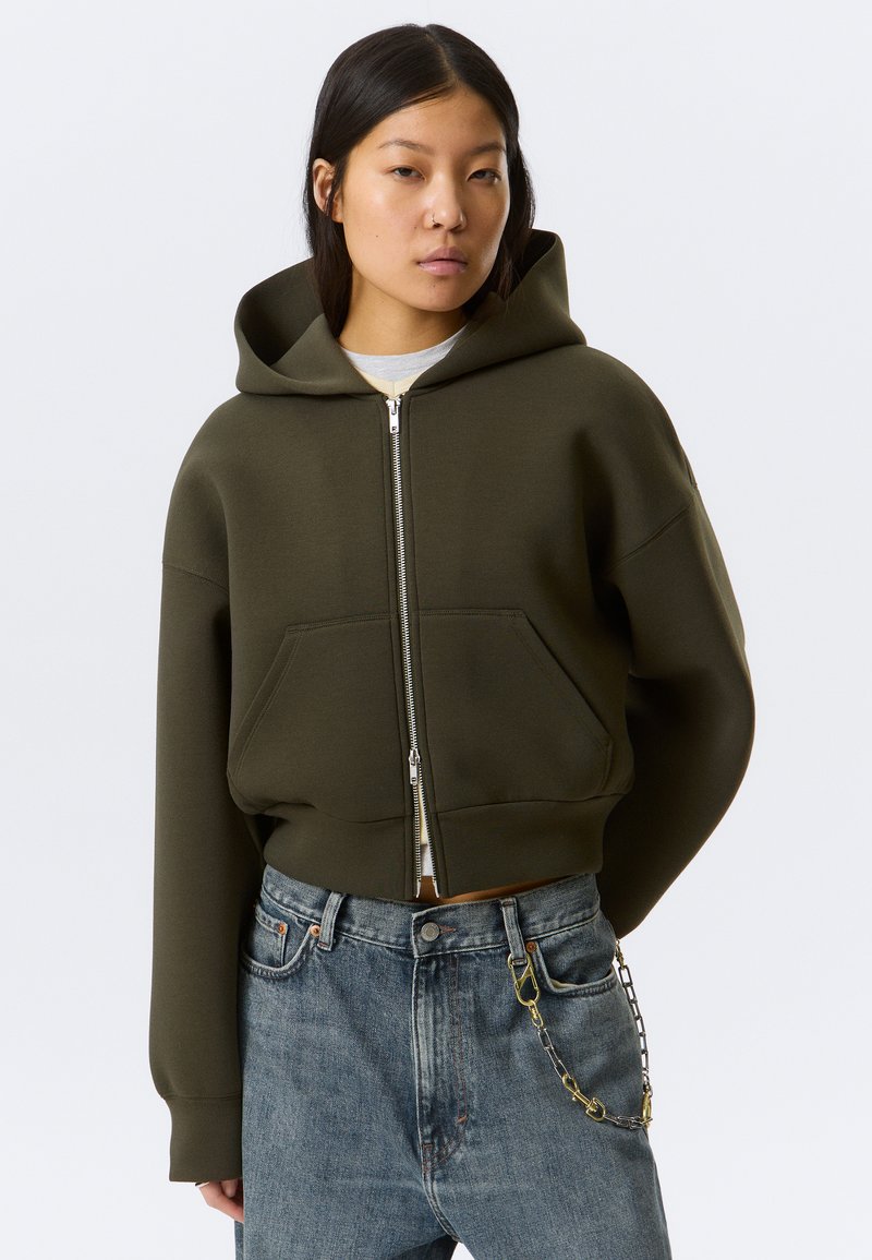 Νεαρή γυναίκα που φοράει μια σκούρα πράσινη cropped ζακέτα με φερμουάρ και μπλε τζιν με μεταλλική αλυσίδα στην δεξιά τσέπη, στέκεται μπροστά σε ένα απλό φόντο.