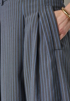 Roka atpūšas zilas un pelēkas vertikāli svītrainas plīsejuma svārka kabatā, uz plaukstas zelta rokassprādze.