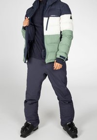 Puffy, color-blocked jacket in navy, white, and green with a zippered pocket and hood. Worn with dark pants and black ski boots.