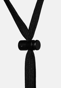 Schwarzes Stoffkabel mit einer strukturierten Oberfläche, zu einer V-Form verdreht und in der Mitte von einem zylindrischen schwarzen Verschluss gesichert.