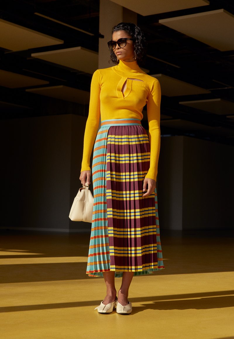 Haut à manches longues jaune avec un trou clé; jupe mi-longue plissée à motifs à carreaux multicolores avec des rayures bleu, bordeaux et jaune; sac à main blanc.