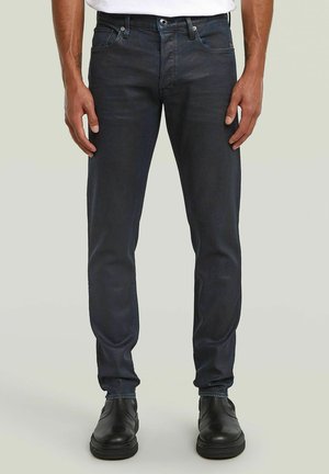 Homme portant un jean slim foncé, des chaussures en cuir noires et une chemise blanche, debout devant un fond clair uni.