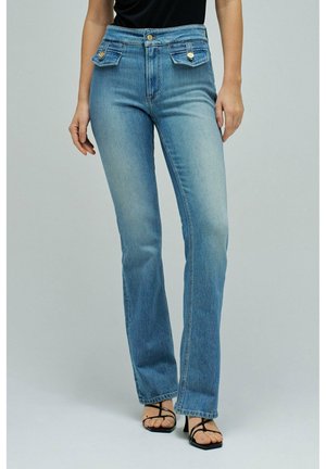 Mujer con vaqueros azules de cintura alta con bolsillos con solapa abotonada, combinados con sandalias negras de tacón con tiras.