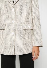 Krémový strukturovaný blejzr s tkaným vzorem, s dvěma předními kapsami na klopu, třemi knoflíky a dlouhými rukávy. Nosi se přes černé kalhoty.