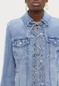 Lys blå denimjakke med snøring i front, utstyrt med gullfarget detaljer, to brystlommer og krage.