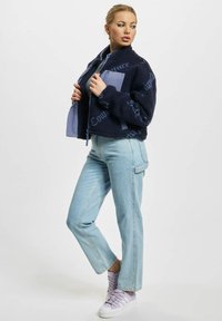 Ženska nosi mornarsko modro flis jakno s svetlo modrimi žepki na prsih, svetlo modre kavbojke in lila superge, stoji z eno nogo upognjeno in gleda ob strani.